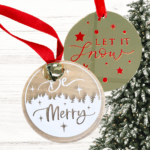 two acrylic ornaments with christmas sayings over a christmas tree